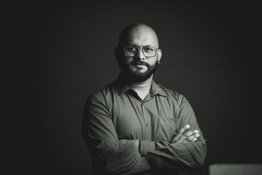Monochrome studio portrait of a confident South Asian man with glasses and a beard, arms crossed.