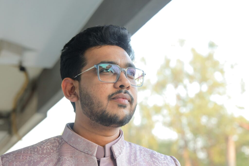 A stylish man wearing glasses and traditional attire, standing outdoors during the day.